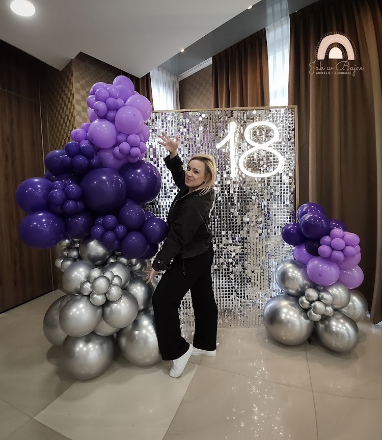 Aranżacja z fioletowych i srebrnych balonów na tle błyszczącej ścianki z neonowym napisem '18', z pozującą kobietą w czarnym stroju.