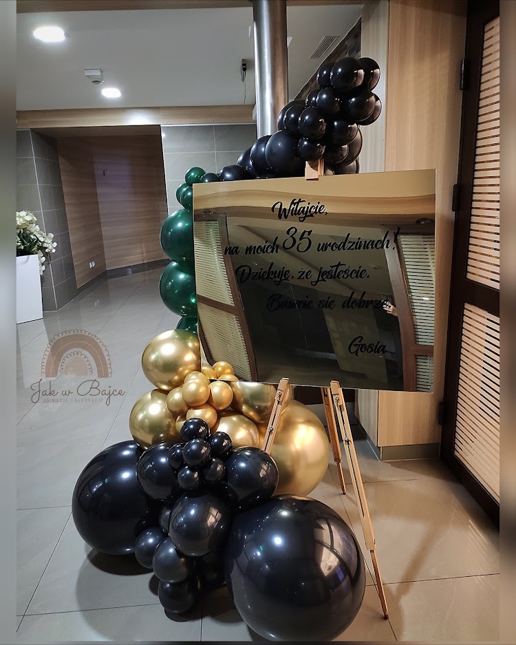 Dekoracja urodzinowa z czarnymi, złotymi i zielonymi balonami, oraz lustrem z napisem powitalnym na drewnianym stojaku, w tle widoczna ściana i podłoga z płytek.
