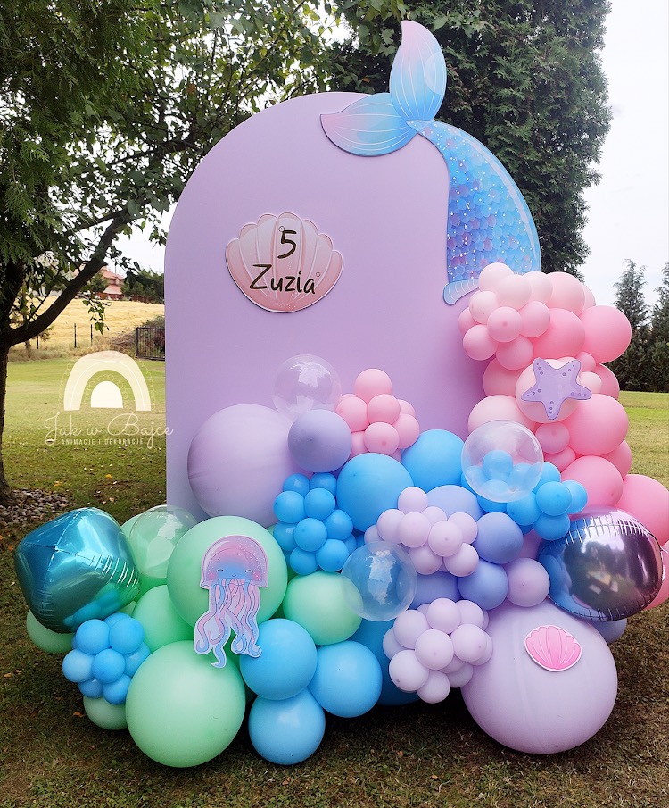 Dekoracja urodzinowa w stylu syreny: pastelowe balony w odcieniach błękitu, fioletu i różu tworzą kompozycję z sylwetką ogona syreny, muszelką z imieniem Zuza i numerem 5, oraz naklejką z meduzą...