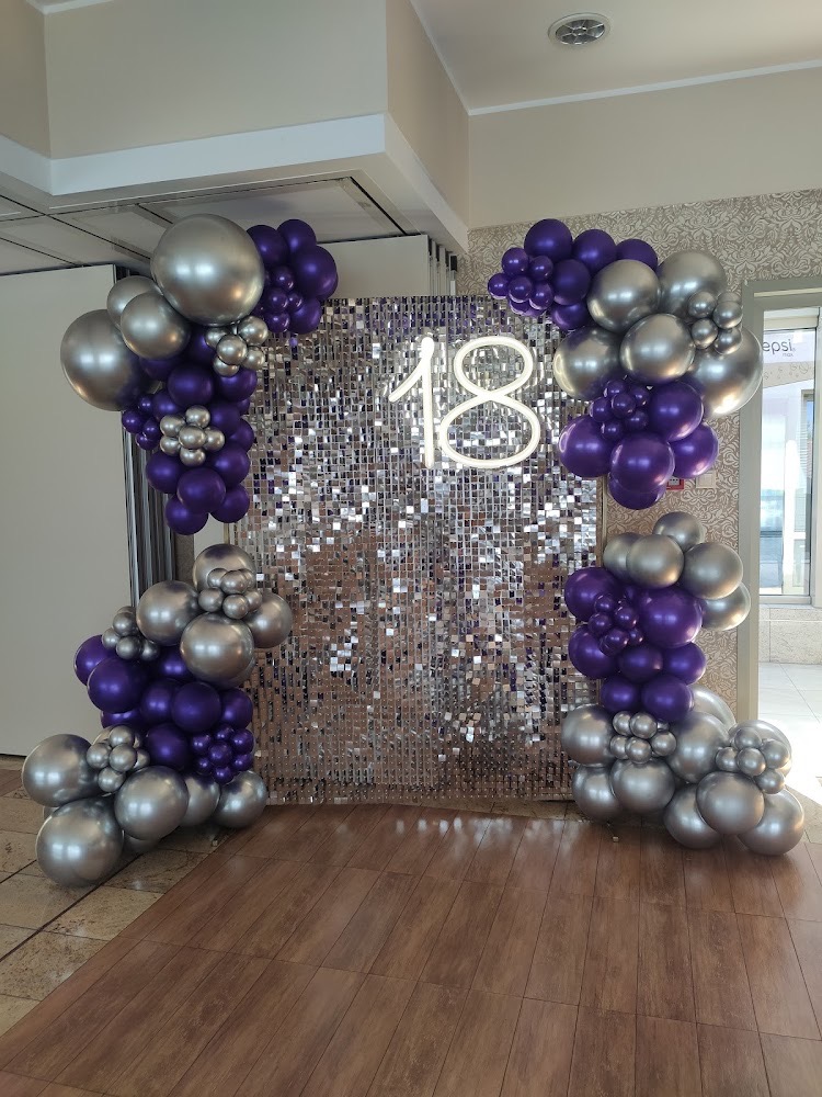 Dekoracja na osiemnaste urodziny: błyszcząca ścianka, neonowy napis '18' oraz girlandy z fioletowych i srebrnych balonów.
