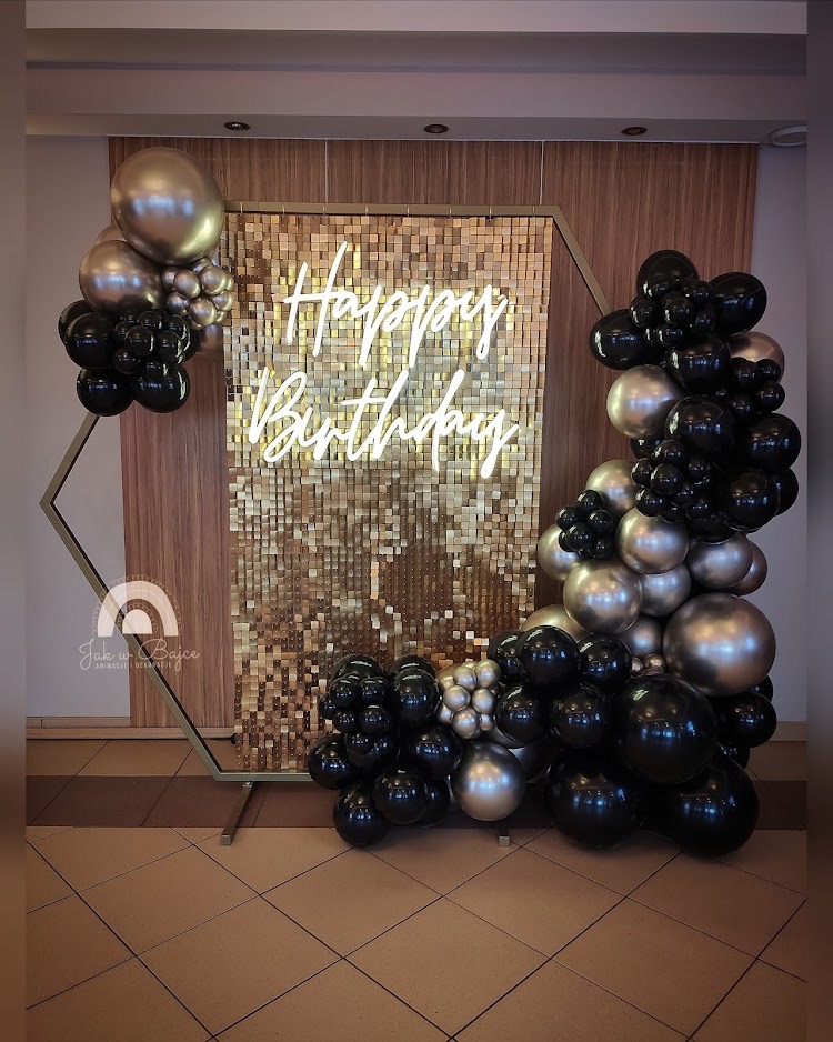 Dekoracja urodzinowa z heksagonalną ramą, złotym błyszczącym tłem, neonowym napisem 'Happy Birthday' oraz girlandą z czarnych i srebrnych balonów.