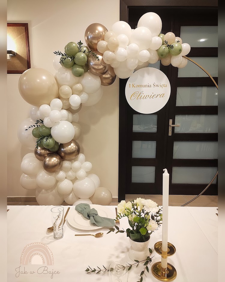 Dekoracja stołu na Pierwszą Komunię Świętą z balonami w odcieniach bieli, beżu, złota i zieleni, z tabliczką z imieniem dziecka, elegancka zastawa stołowa i świeże kwiaty.