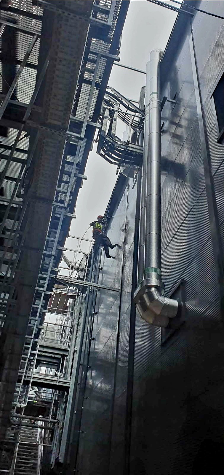 Pracownik na linach alpinistycznych spryskuje wodą metalową elewację przemysłowego budynku, widoczne rury wentylacyjne i metalowe konstrukcje. Ujęcie z dołu.