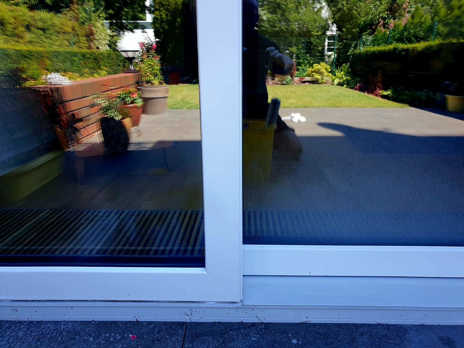 Pracownik z wiadrem i myjką czyści duże przesuwne okno tarasowe, odbicie ogrodu w szybie.