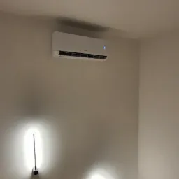 Biała klimatyzacja ścienna włączona, zamontowana nad schodami, z dwoma podłużnymi lampami po bokach, oświetlającymi klatkę schodową.