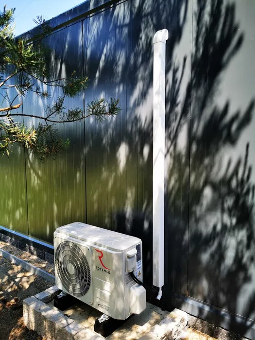 Zewnętrzna jednostka klimatyzacji Rotenso zamontowana na betonowych bloczkach przy metalowym panelu ściennym z cieniem drzewa.