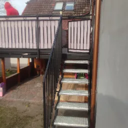 Zewnętrzne schody metalowe z kratowymi stopniami prowadzące na balkon z balustradą, widok z boku budynku.