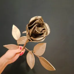 Ręcznie wykonana, metalowa róża w odcieniach złota, trzymana w dłoni z czerwonym manicure na tle ciemnej ściany.