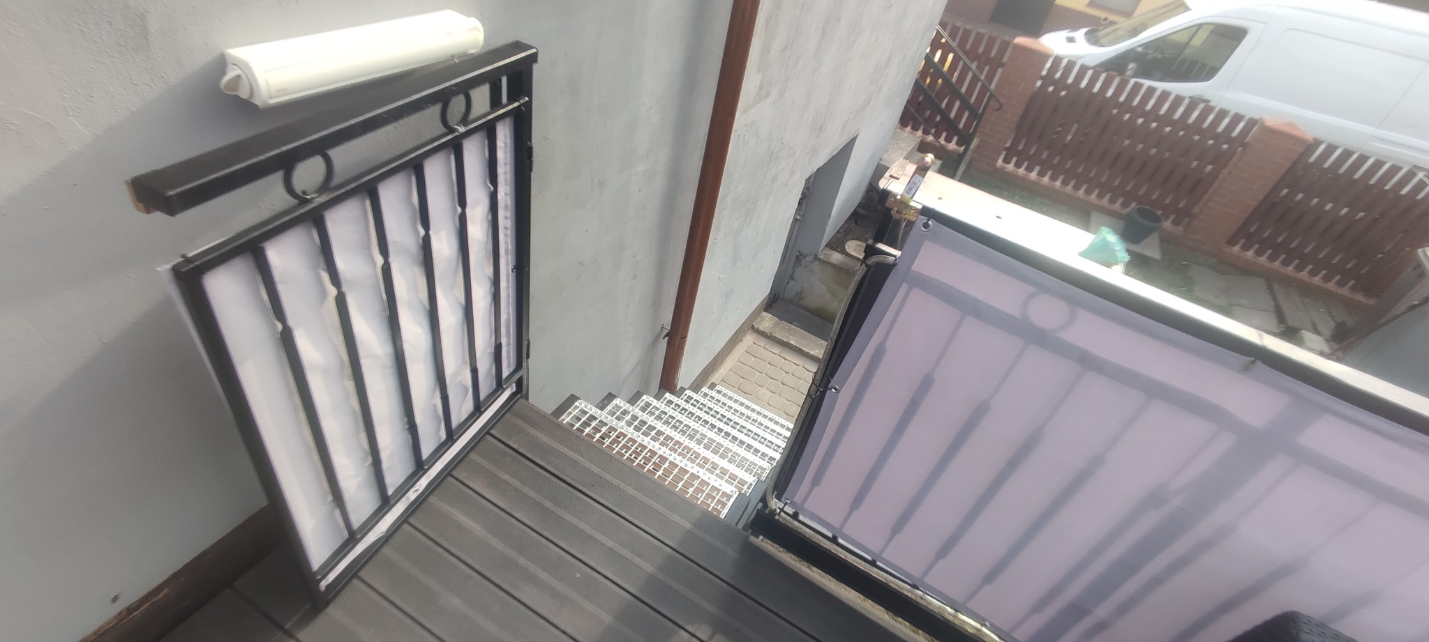 Metalowa balustrada z czarnymi elementami i białym wypełnieniem, schody z metalowej siatki i drewnopodobnych desek, widok z góry na podwórko z samochodem dostawczym w tle.