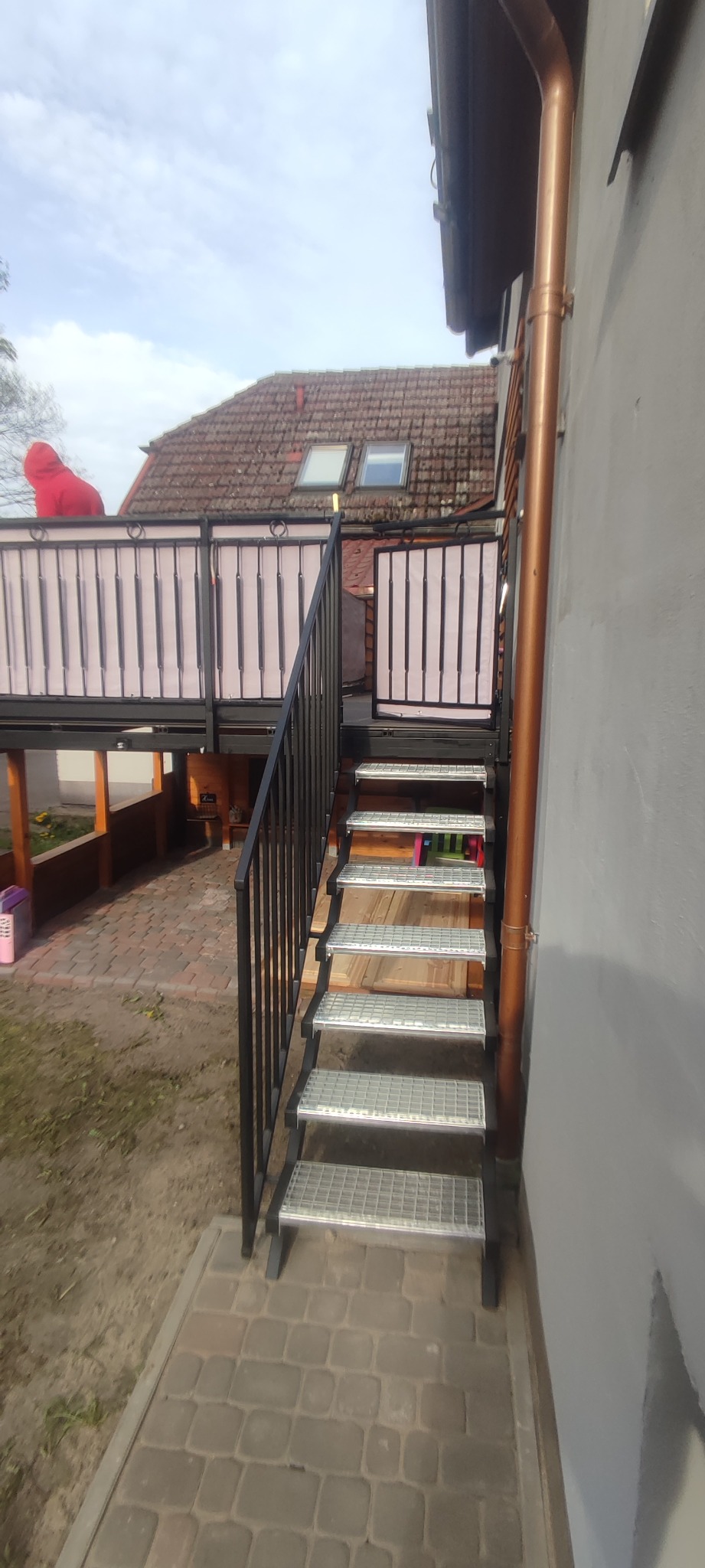 Zewnętrzne schody metalowe z kratowymi stopniami prowadzące na balkon z balustradą, widok z boku budynku.