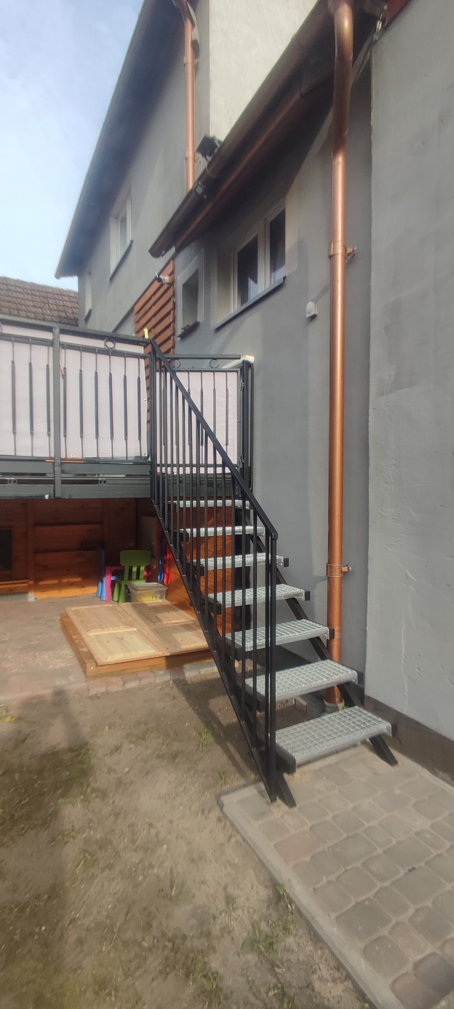 Zewnętrzne schody metalowe z ażurowymi stopniami i czarną balustradą prowadzące na balkon z białą osłoną, widoczne rury spustowe, szara elewacja budynku i częściowo widoczny drewniany domek.