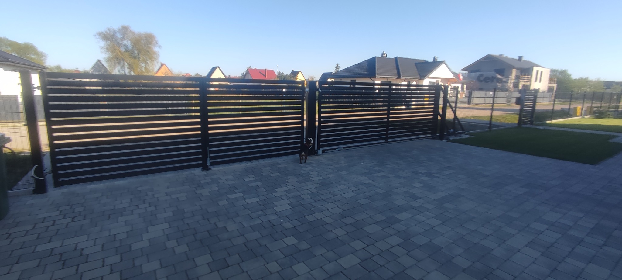 Nowoczesna, pozioma brama wjazdowa z czarnych profili, z małym psem stojącym pośrodku, na tle kostki brukowej i domów jednorodzinnych.