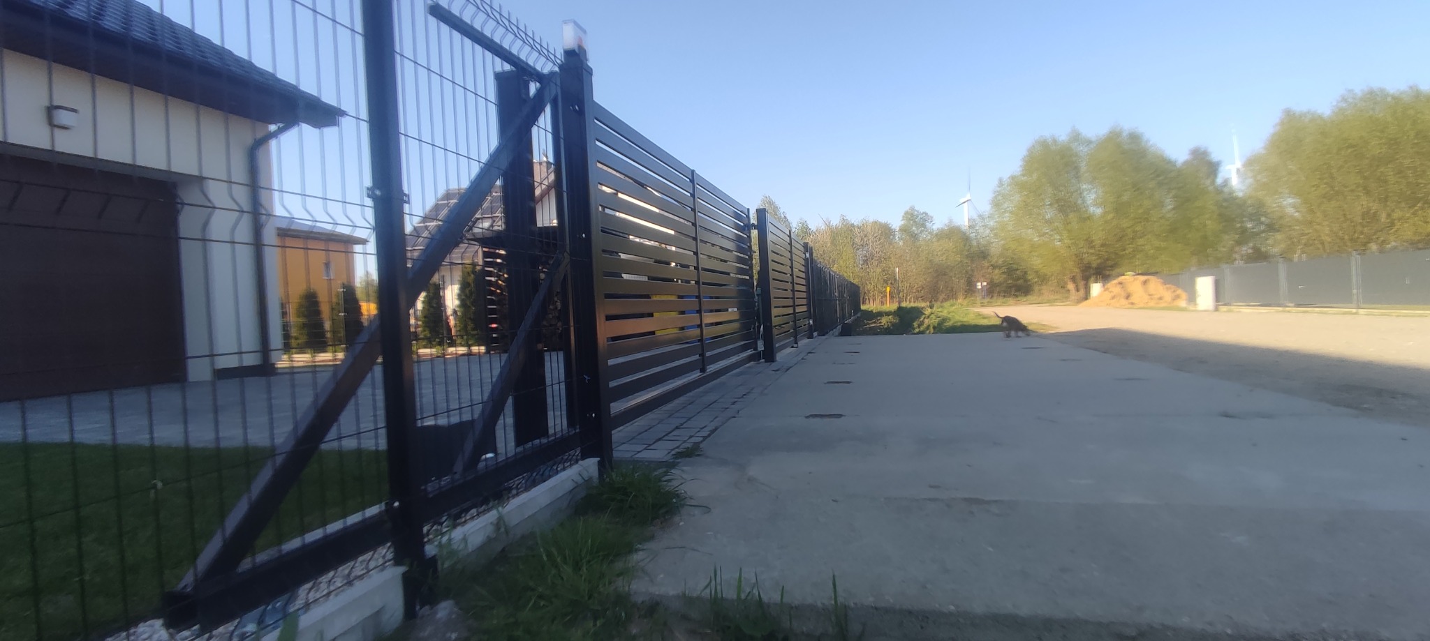 Nowoczesna brama posesyjna z poziomymi panelami, czarna, z widocznym fragmentem ogrodzenia panelowego, na tle domu jednorodzinnego i drzew.