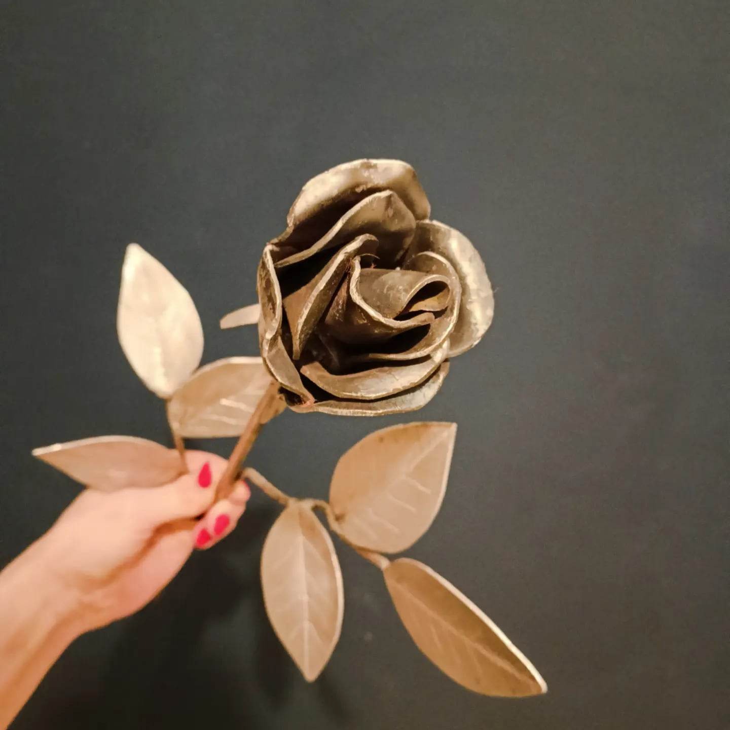 Ręcznie wykonana, metalowa róża w odcieniach złota, trzymana w dłoni z czerwonym manicure na tle ciemnej ściany.