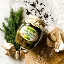 Wiktoria Bęben - Słoik ogórków kiszonych marki Waldi-Ben na drewnianej desce z czosnkiem, koperkiem, solą i ziarnami pieprzu na pomiętym białym papierze.