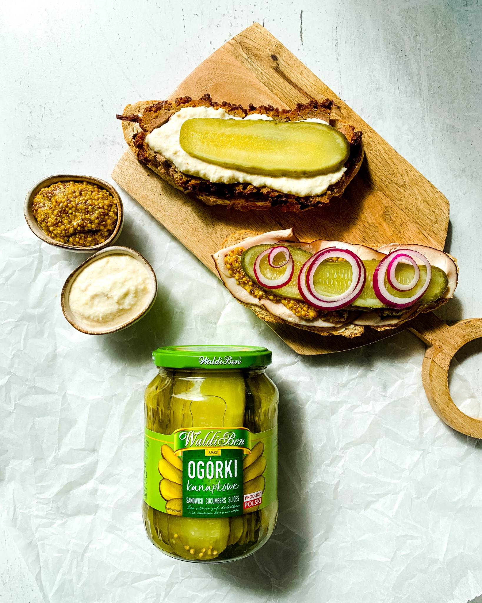 Dwa otwarte tosty na drewnianych deskach, jeden z kremowym serem i plasterkiem ogórka, drugi z szynką, musztardą, ogórkiem i krążkami czerwonej cebuli, obok miseczki z musztardą ziarnistą i sosem...