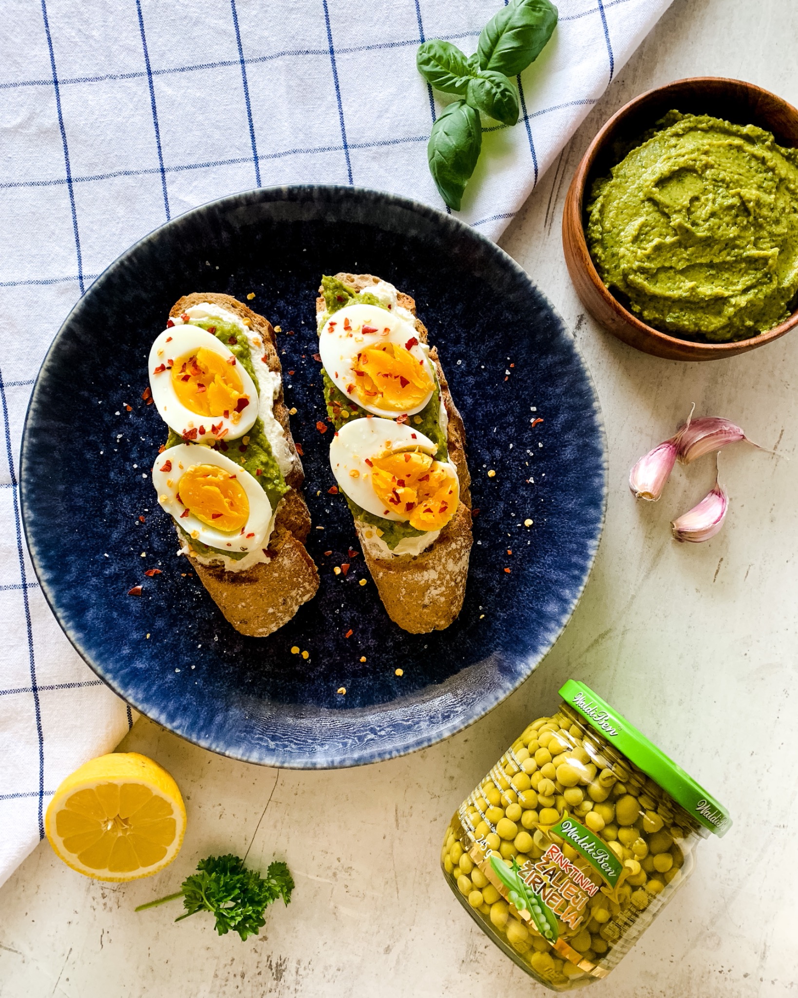 Dwa tosty z chleba razowego z pastą z zielonego groszku, białym serem i jajkiem na twardo, posypane płatkami chili, na talerzu obok miseczki z pastą, ząbków czosnku, cytryny, gałązki bazylii...