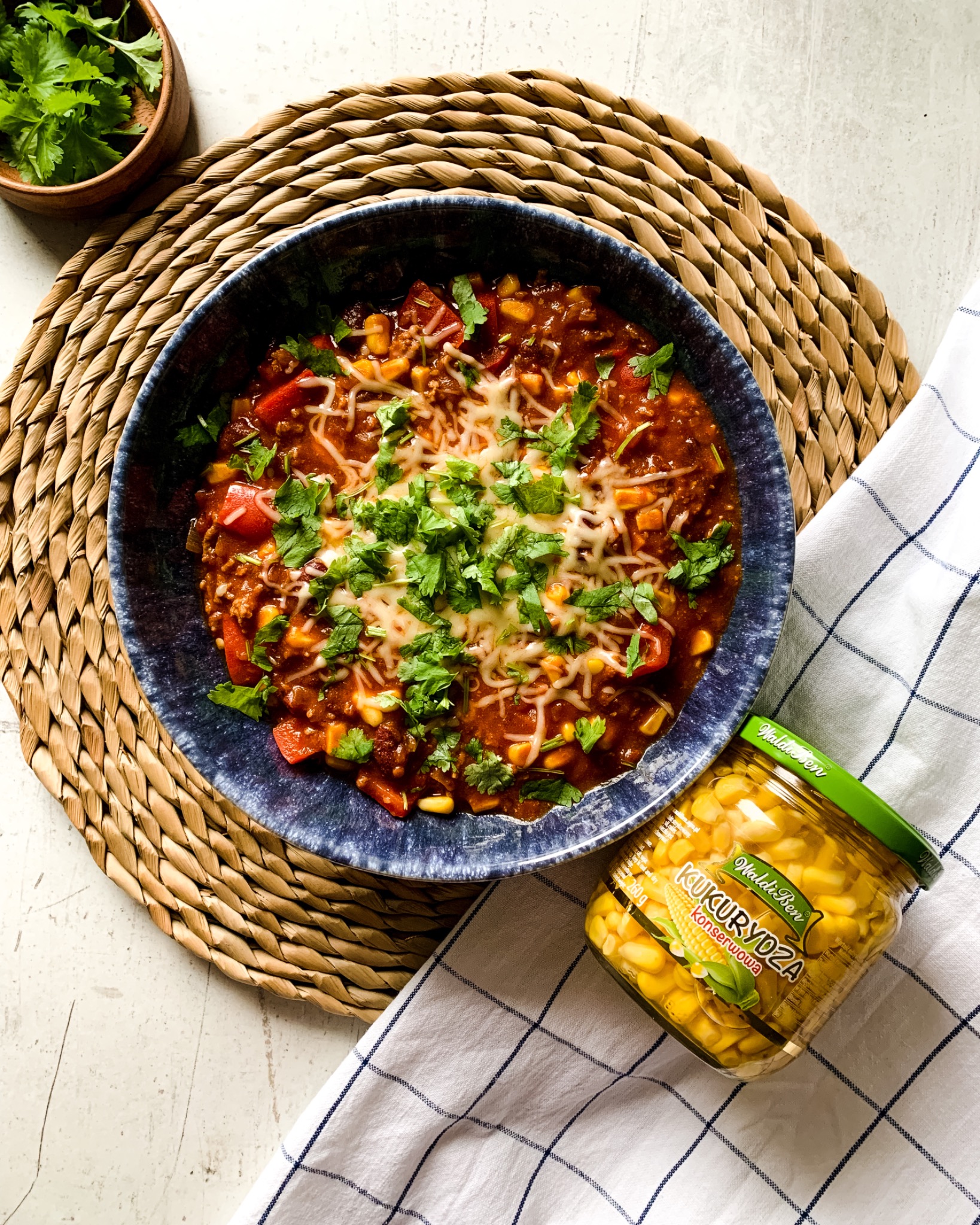 Chili con carne z kukurydzą, papryką i serem, posypane świeżą kolendrą, podane w niebieskiej misce na słomianej podkładce, obok słoik konserwowej kukurydzy WaldiBen na białej ściereczce w niebieską...