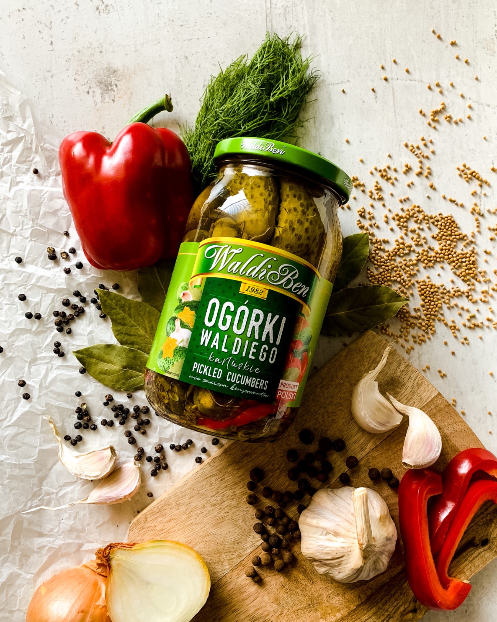 Słoik ogórków konserwowych marki Waldi Ben otoczony świeżymi warzywami i przyprawami, w tym czerwoną papryką, czosnkiem, cebulą, koprem, ziarnami gorczycy i pieprzem, ułożony na drewnianej desce...