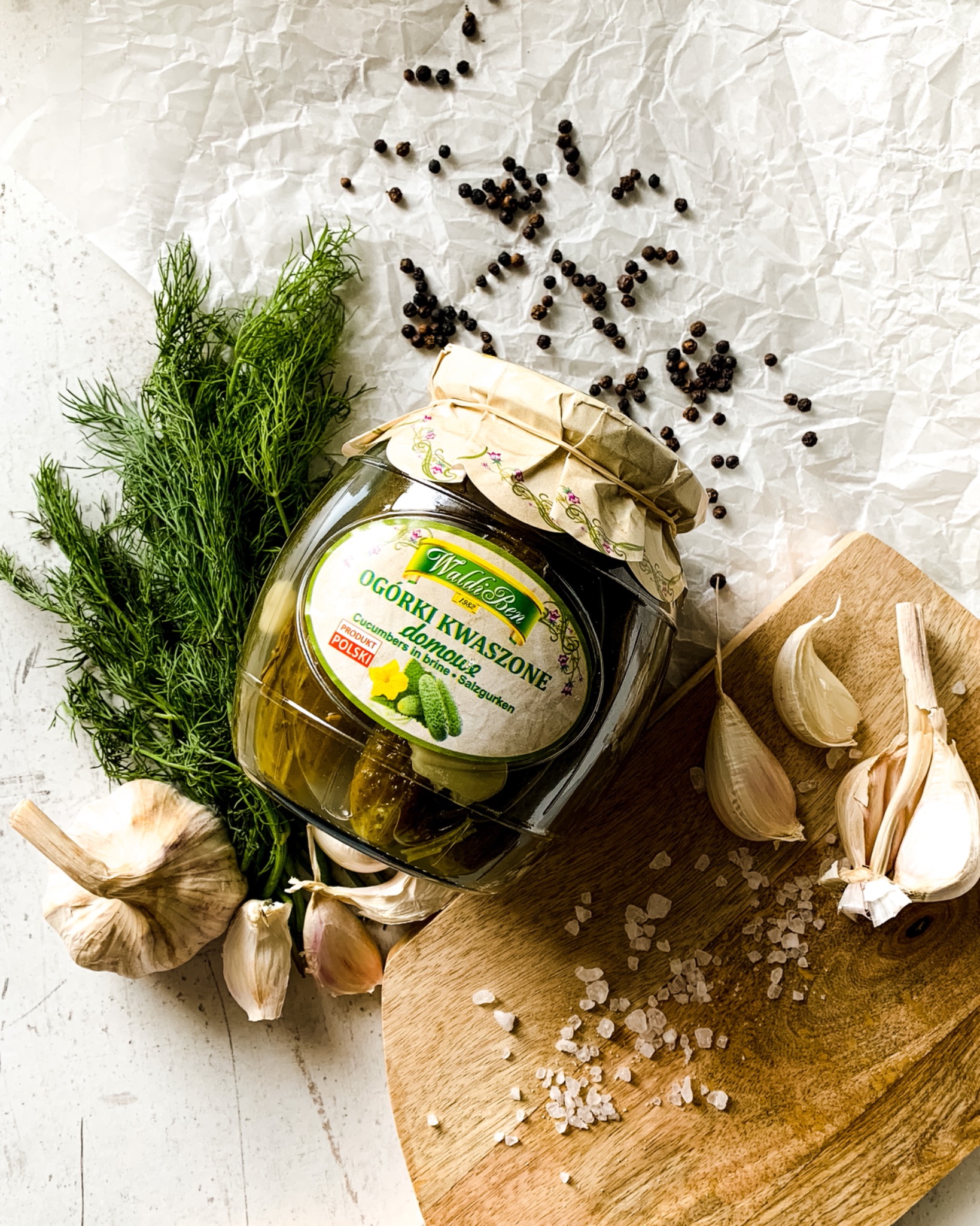 Słoik ogórków kiszonych marki Waldi-Ben na drewnianej desce z czosnkiem, koperkiem, solą i ziarnami pieprzu na pomiętym białym papierze.