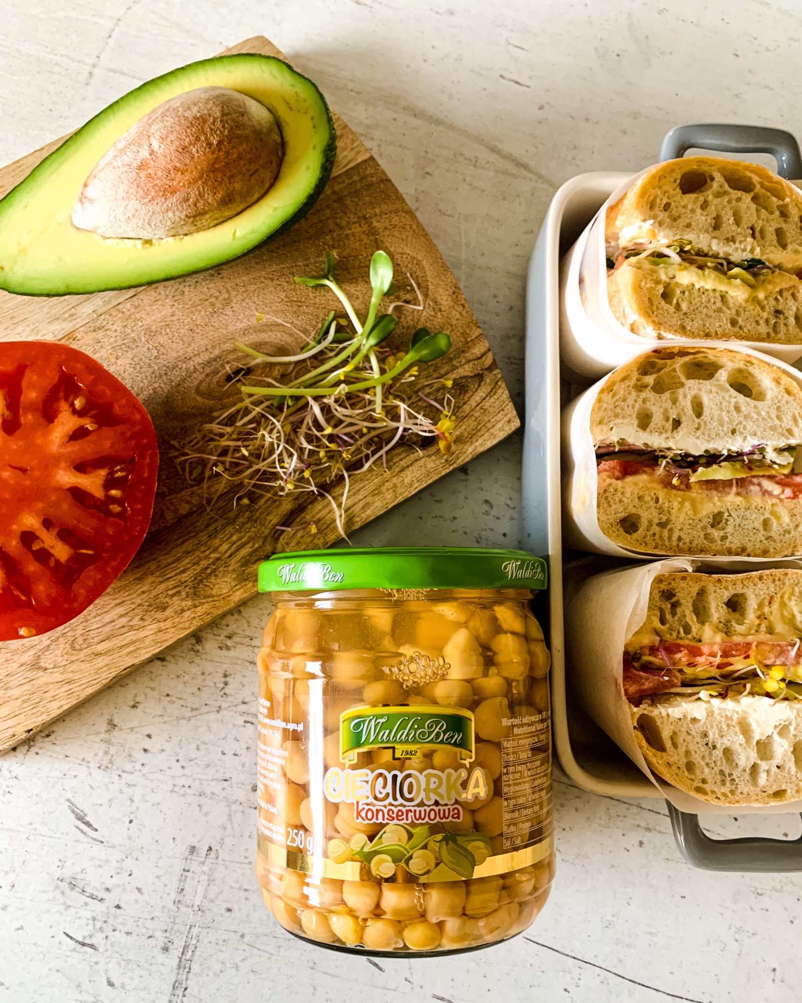 Kompozycja kulinarna z przekrojem awokado, pomidora, kiełków, słoikiem ciecierzycy konserwowej Waldi Ben i trzema kanapkami w papierze, umieszczonymi w szarym pojemniku.