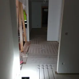 Wnętrze w trakcie montażu ogrzewania podłogowego, widoczna rozłożona instalacja na podłodze, lampa budowlana oświetlająca fragment pomieszczenia, w tle drzwi do kolejnego pokoju.