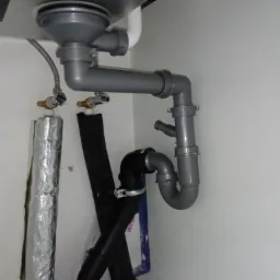 Szare i czarne rury odpływowe pod zlewem, widoczne zawory dopływu wody z izolacją termiczną, ujęcie z dołu.