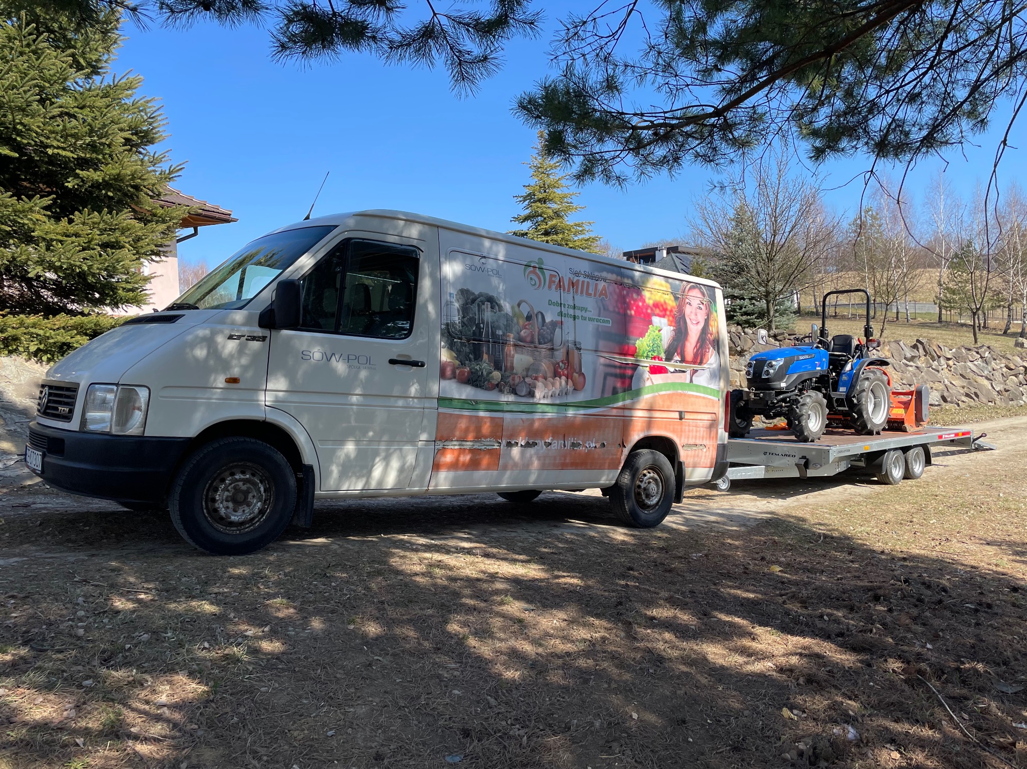 Biały bus z reklamą sieci sklepów Familia ciągnie przyczepę z niebieskim traktorem na tle drzew i błękitnego nieba.