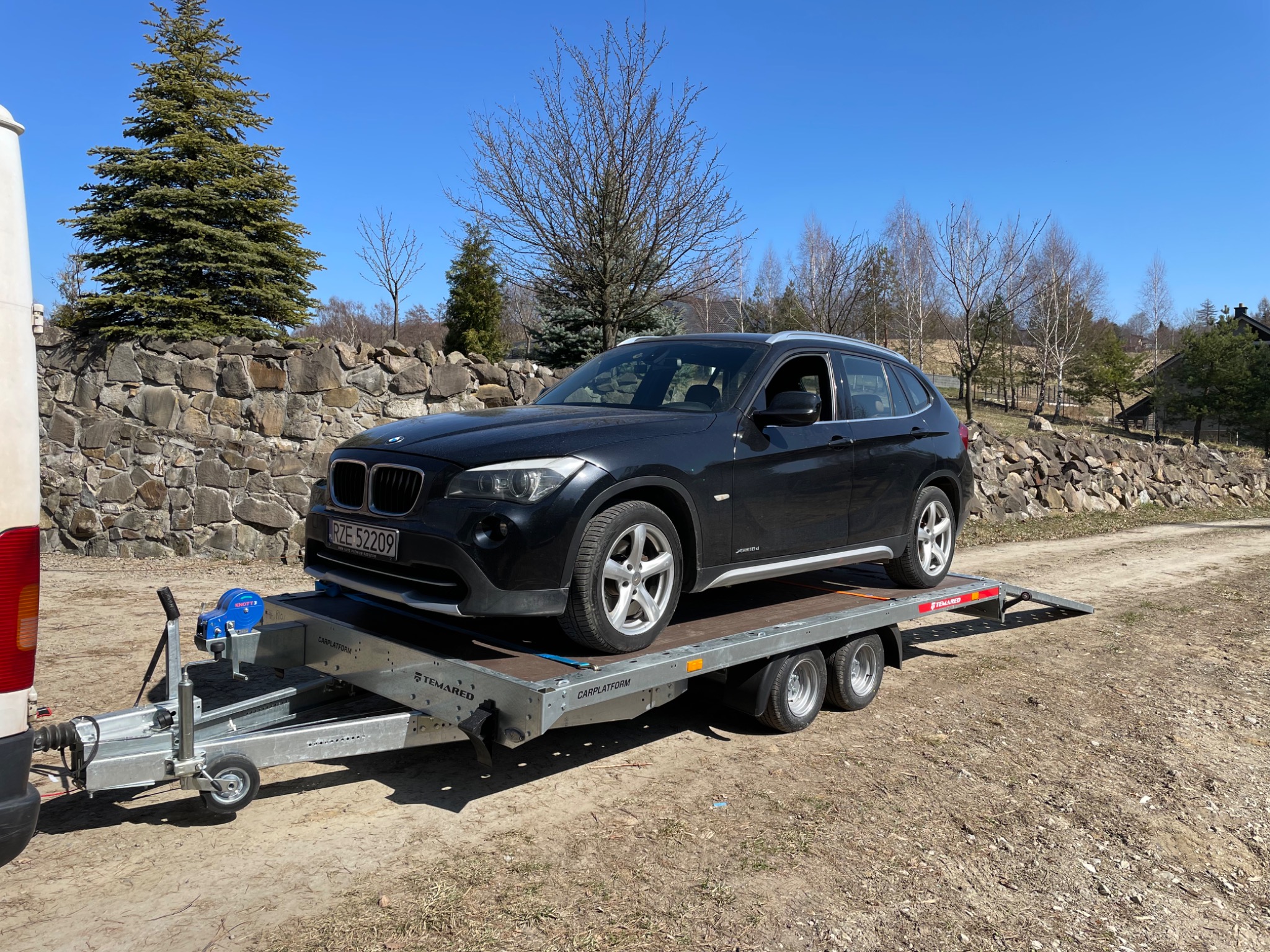 Czarny samochód BMW X1 na platformie przyczepy samochodowej Temared Carplatform, gotowy do transportu, z widocznym fragmentem białego busa i kamiennym murem w tle.