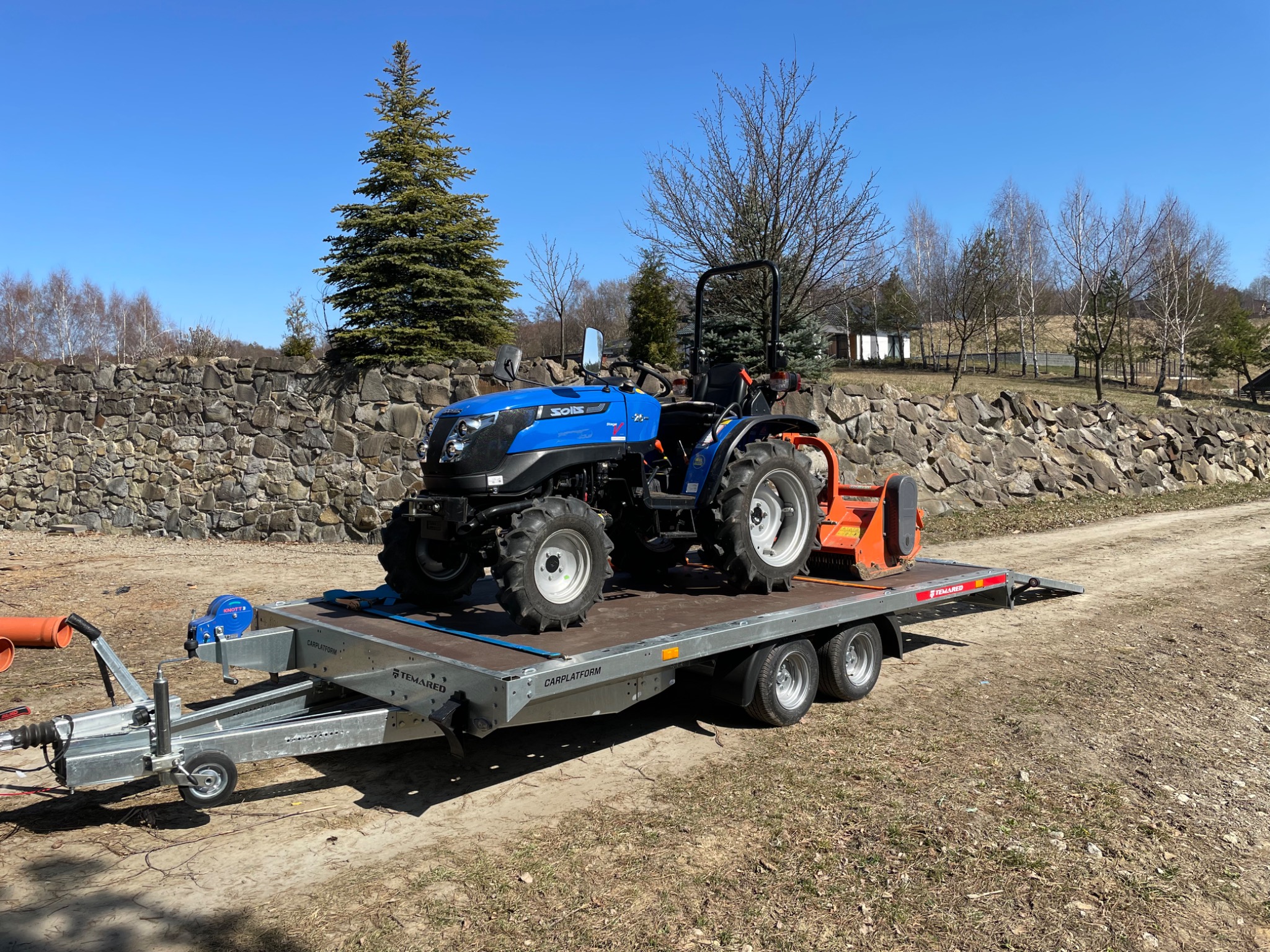 Niebieski ciągnik Solis z pomarańczowym pługiem wirnikowym zamontowanym z tyłu, umieszczony na platformie transportowej Temared, na tle kamiennego muru i drzew w bezlistnym lesie.