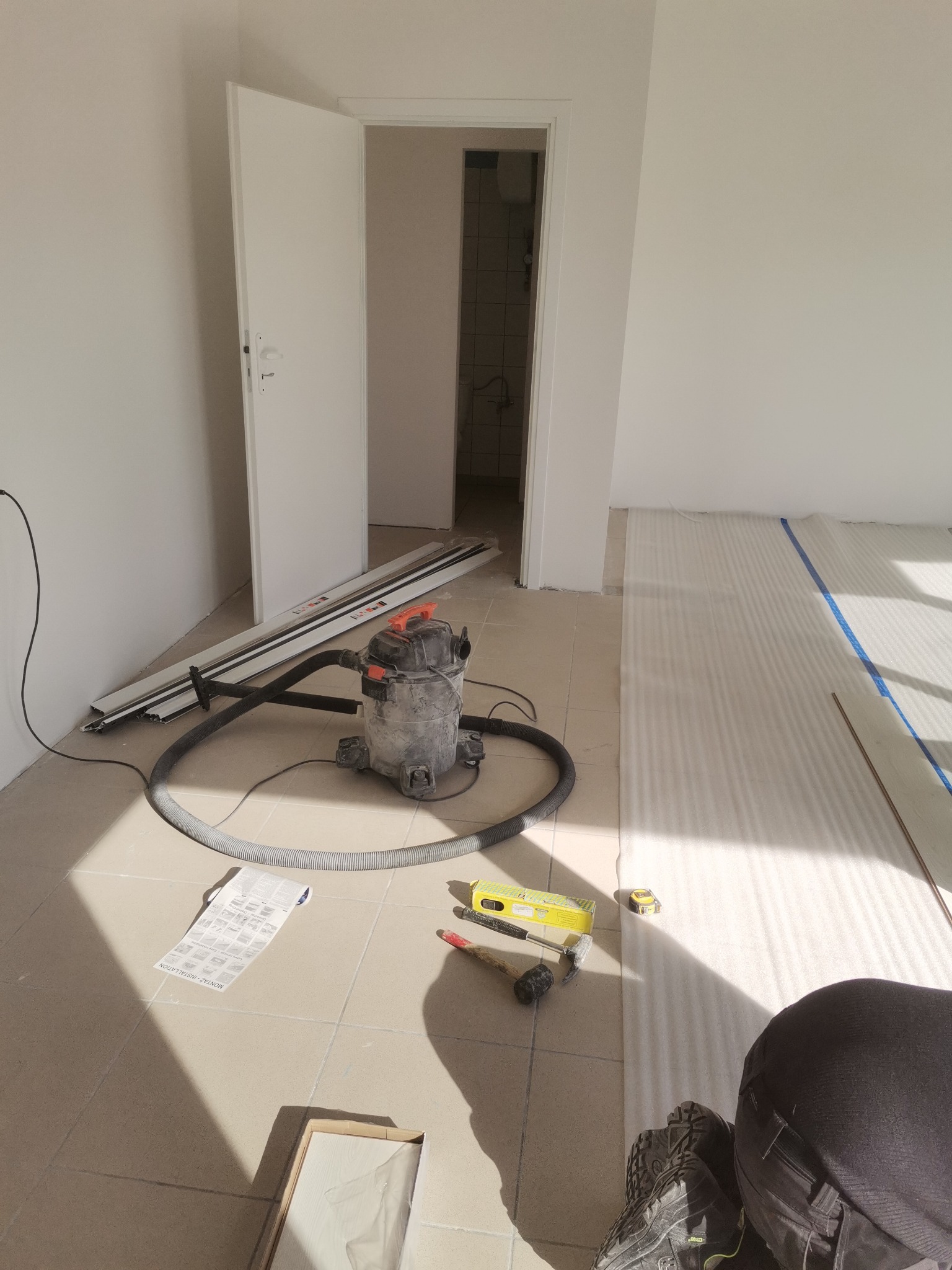 Przygotowanie do montażu paneli podłogowych: rozłożona pianka izolacyjna, narzędzia (poziomica, młotek, miarka), odkurzacz przemysłowy i listwy przypodłogowe w jasnym, remontowanym pomieszczeniu...