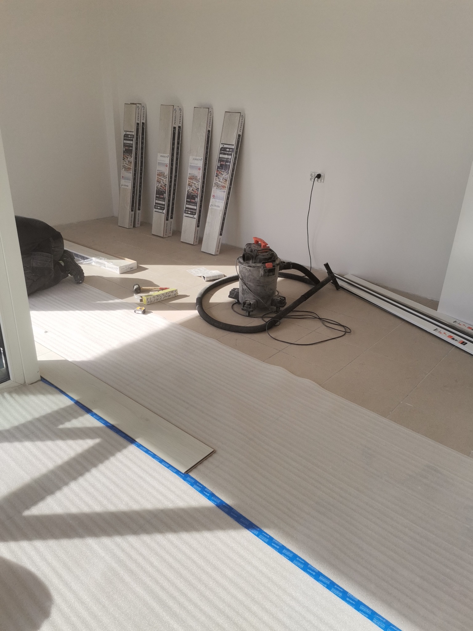 Układanie paneli podłogowych: widok na podkład z taśmą, panele, odkurzacz przemysłowy i narzędzia w jasnym pomieszczeniu.