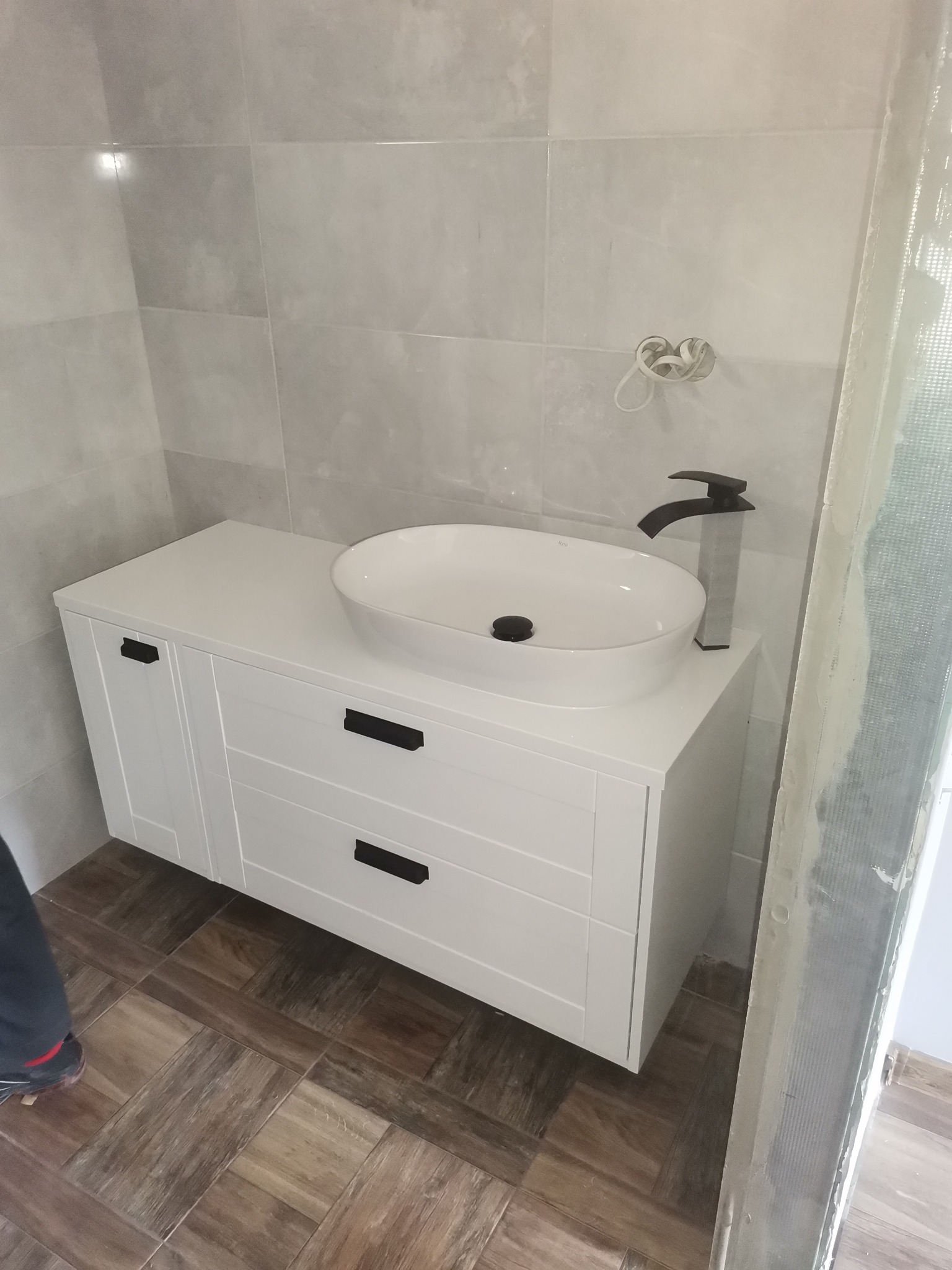 Biała szafka łazienkowa z umywalką nablatową i czarną armaturą, widoczne płytki ścienne w odcieniach szarości i fragment podłogi imitującej drewno, instalacja elektryczna wystająca ze ściany...