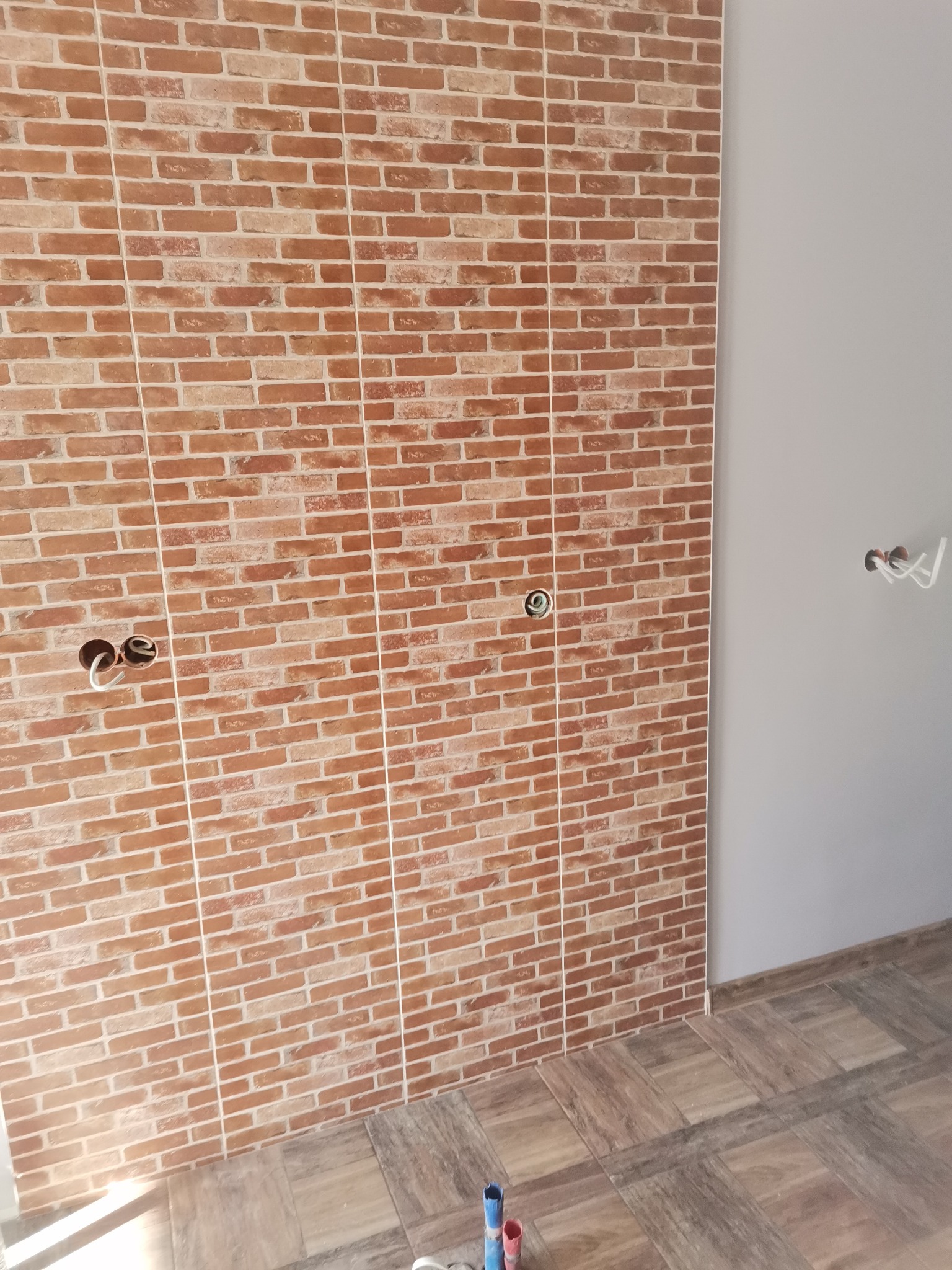 Ściana wyłożona brązowymi płytkami imitującymi cegłę, z widocznymi otworami na instalacje elektryczne i fragmentem szarej ściany obok, oraz podłoga wyłożona płytkami drewnopodobnymi.