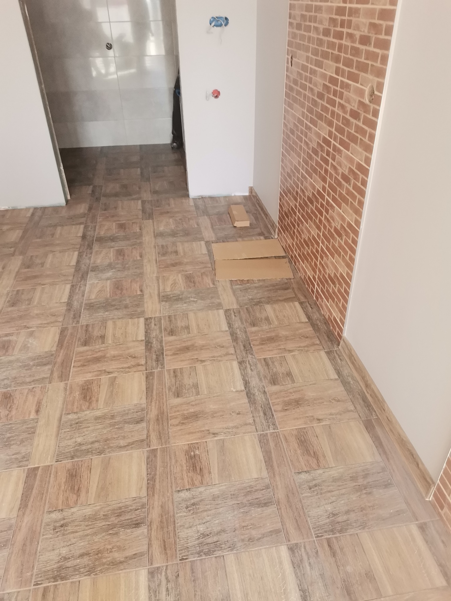 Ułożona podłoga z płytek imitujących drewno w nowym pomieszczeniu, ściana wykończona płytkami imitującymi cegłę, widoczne kartony na podłodze.