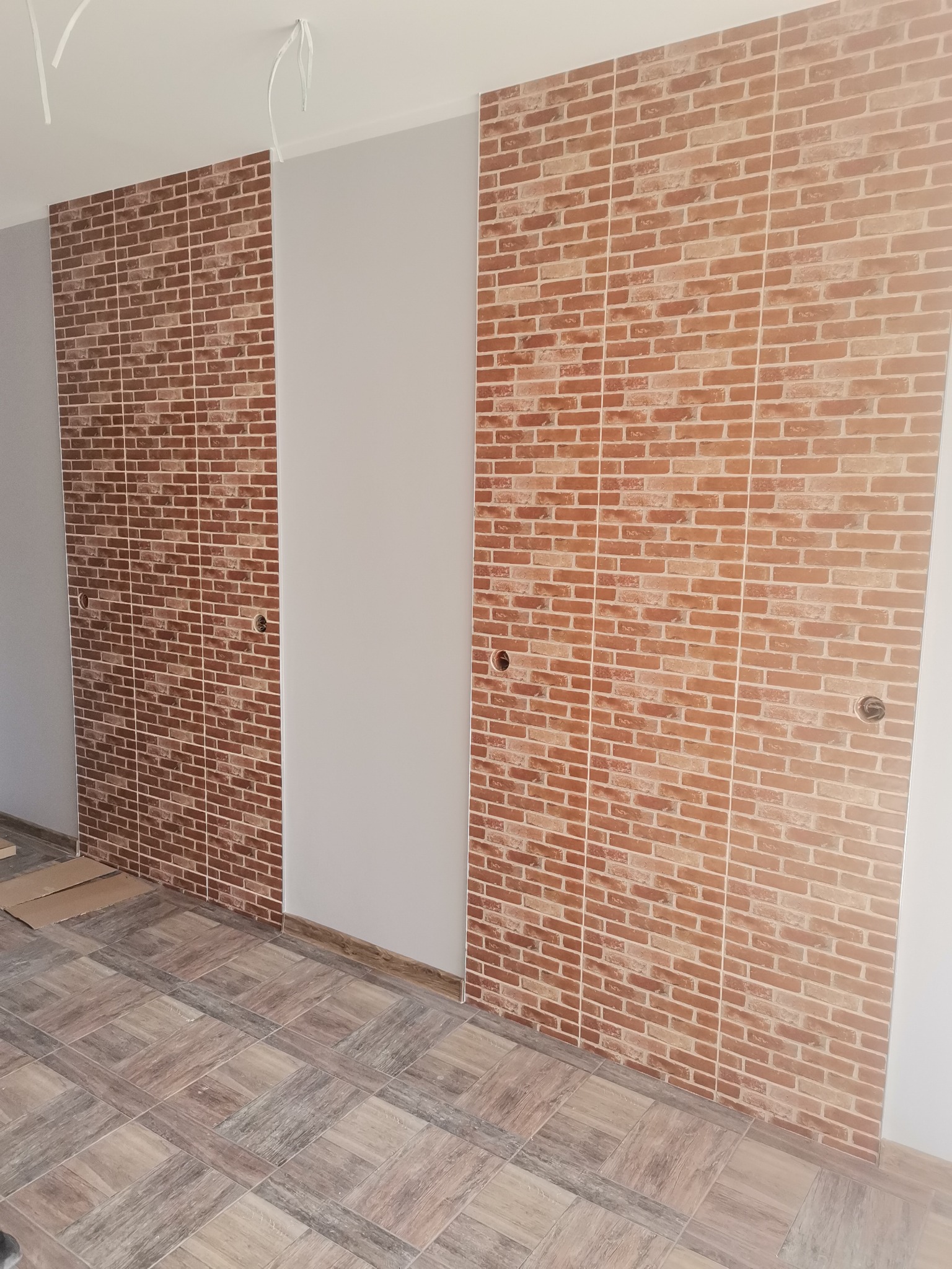 Wykończone wnętrze z brązowymi płytkami imitującymi cegłę na ścianie, dopasowane do podłogi w kwadratowy wzór. Widoczne otwory na gniazdka elektryczne i wystające kable sufitowe.