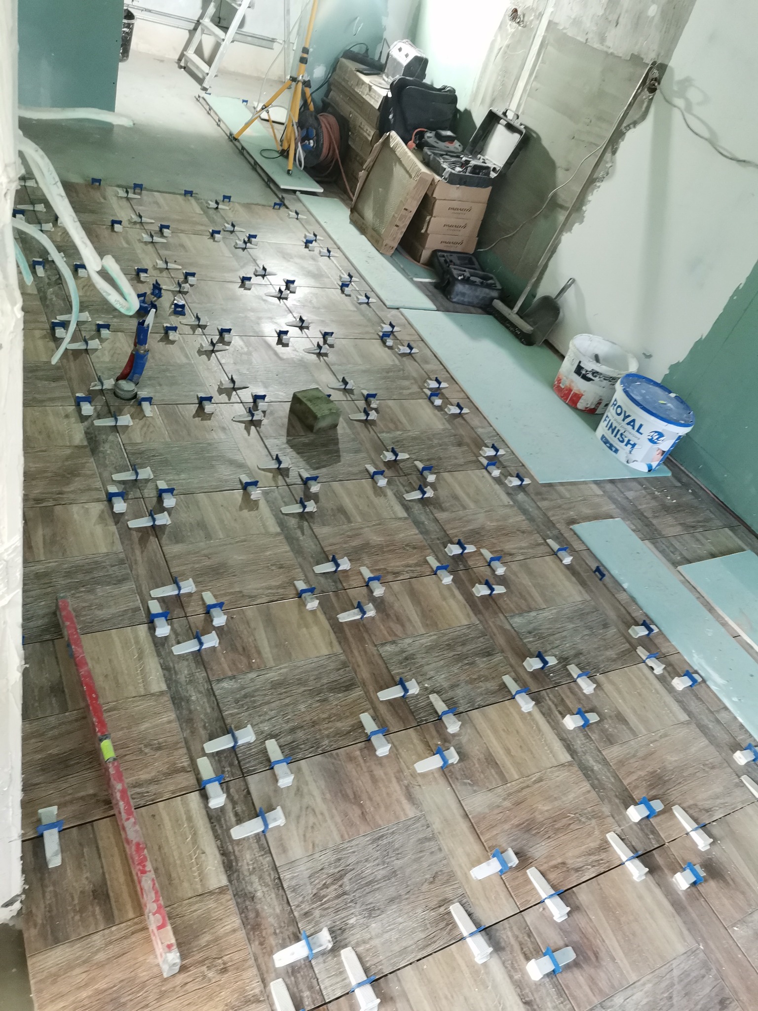 Układanie płytek podłogowych imitujących drewno z użyciem systemu poziomowania; widoczny poziomica, wiadro farby i narzędzia w tle.