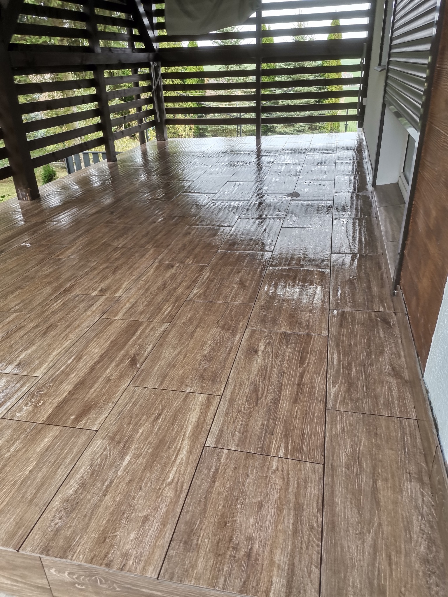 Położona, błyszcząca, drewnopodobna terakota na zadaszonym tarasie z drewnianą balustradą, widoczne odbicia otoczenia w mokrej powierzchni płytek.