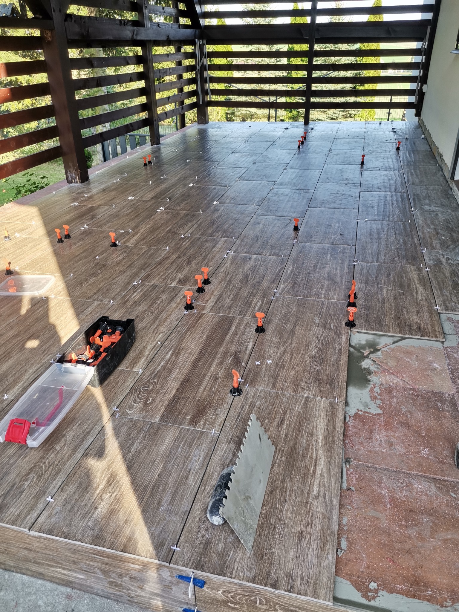 Układanie brązowych płytek imitujących drewno na tarasie z drewnianą konstrukcją z użyciem systemu poziomowania płytek, widoczna paca do nakładania kleju i pojemniki z elementami systemu.