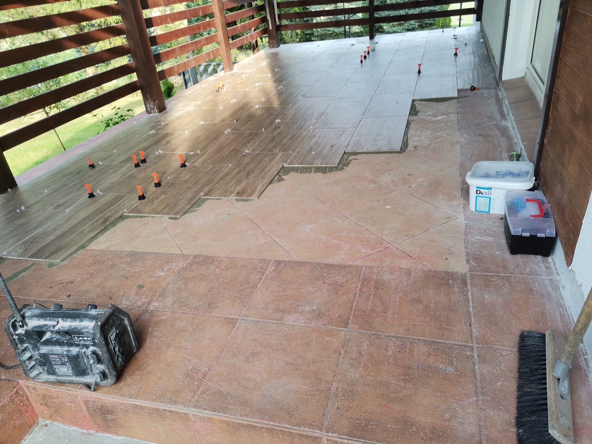 Układanie płytek podłogowych imitujących drewno na tarasie, widoczne narzędzia glazurnicze i poziomica laserowa.