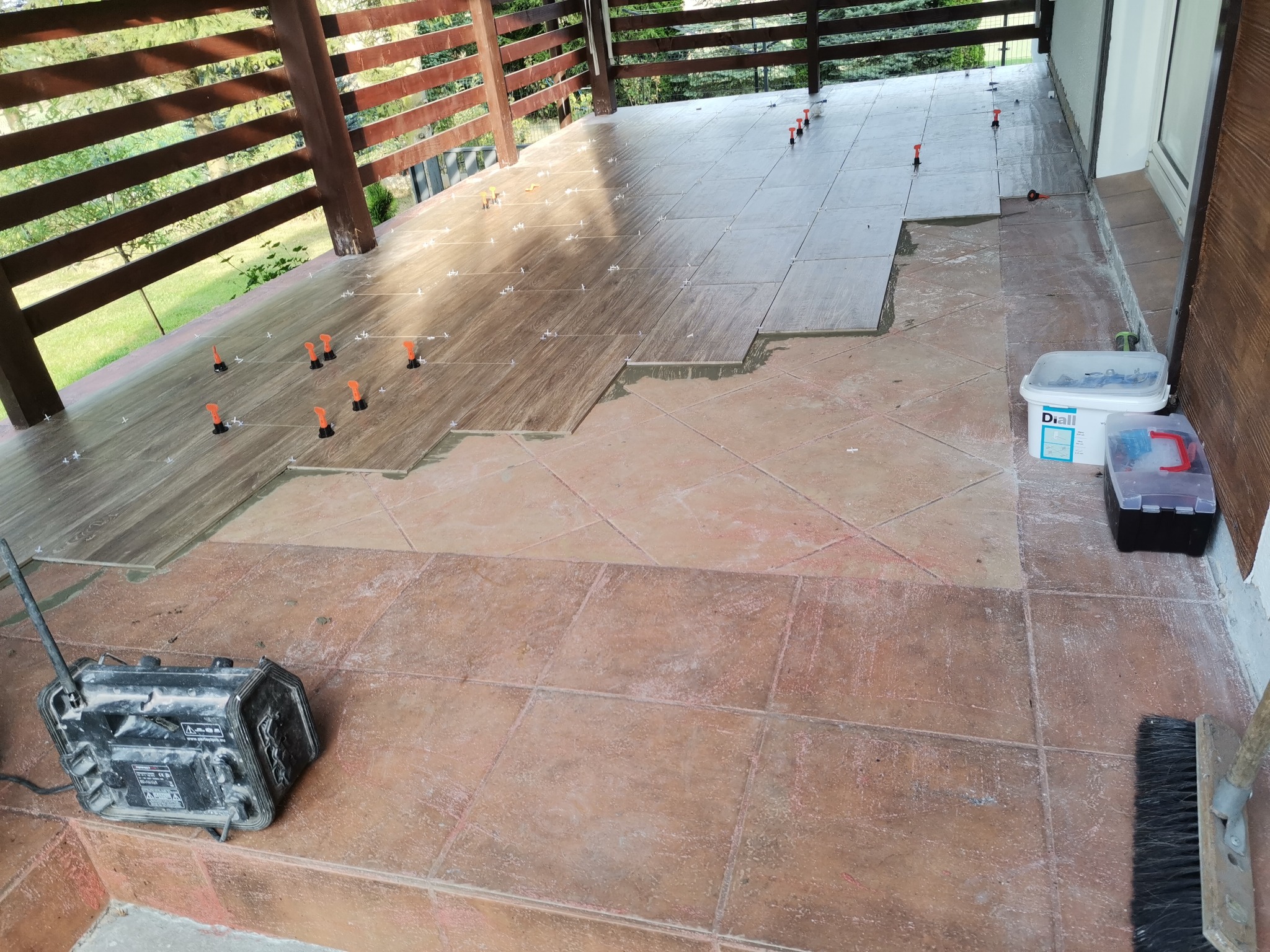 Układanie płytek podłogowych imitujących drewno na tarasie, widoczne poziomice i narzędzia glazurnicze, część powierzchni już wyłożona, druga część z widocznymi starymi płytkami.