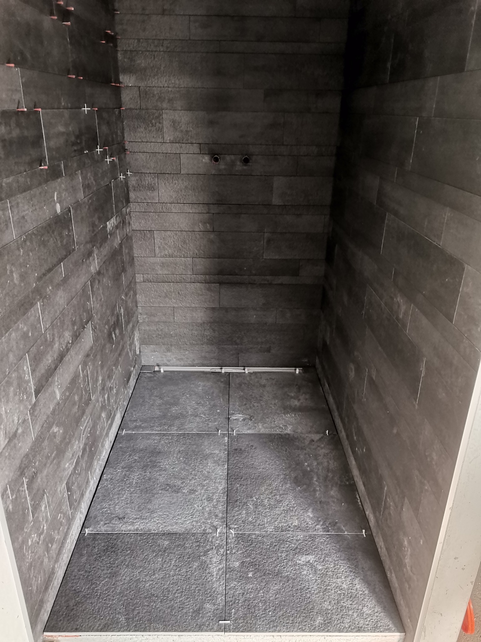Wnętrze kabiny prysznicowej w trakcie układania ciemnoszarych płytek imitujących kamień, widoczne krzyżyki dystansowe i poziomica.