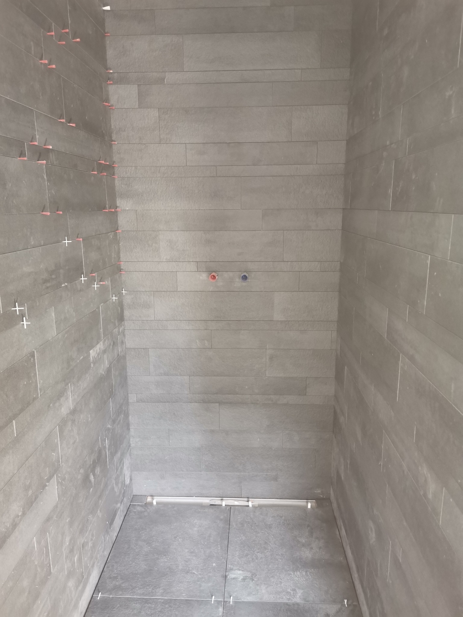 Wnętrze kabiny prysznicowej w trakcie wykańczania, widoczne szare, prostokątne płytki na ścianach i podłodze, krzyżyki dystansowe i poziomica przy odpływie liniowym.
