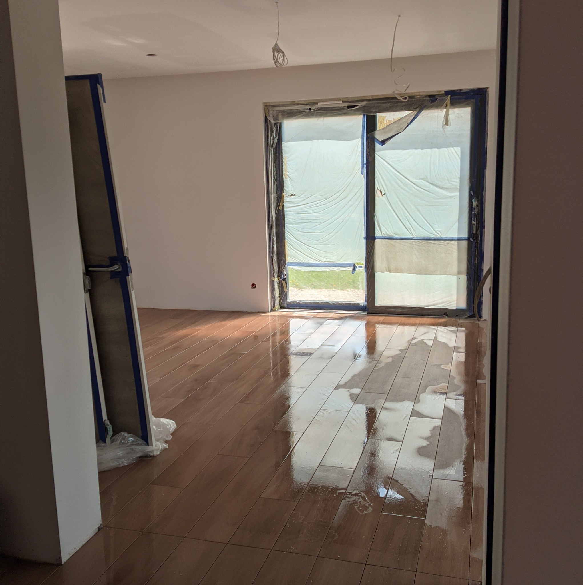 Pokój w trakcie remontu: zabezpieczone okno folią, świeżo położona, mokra podłoga z płytek imitujących drewno, przygotowane do montażu płyty gipsowo-kartonowe