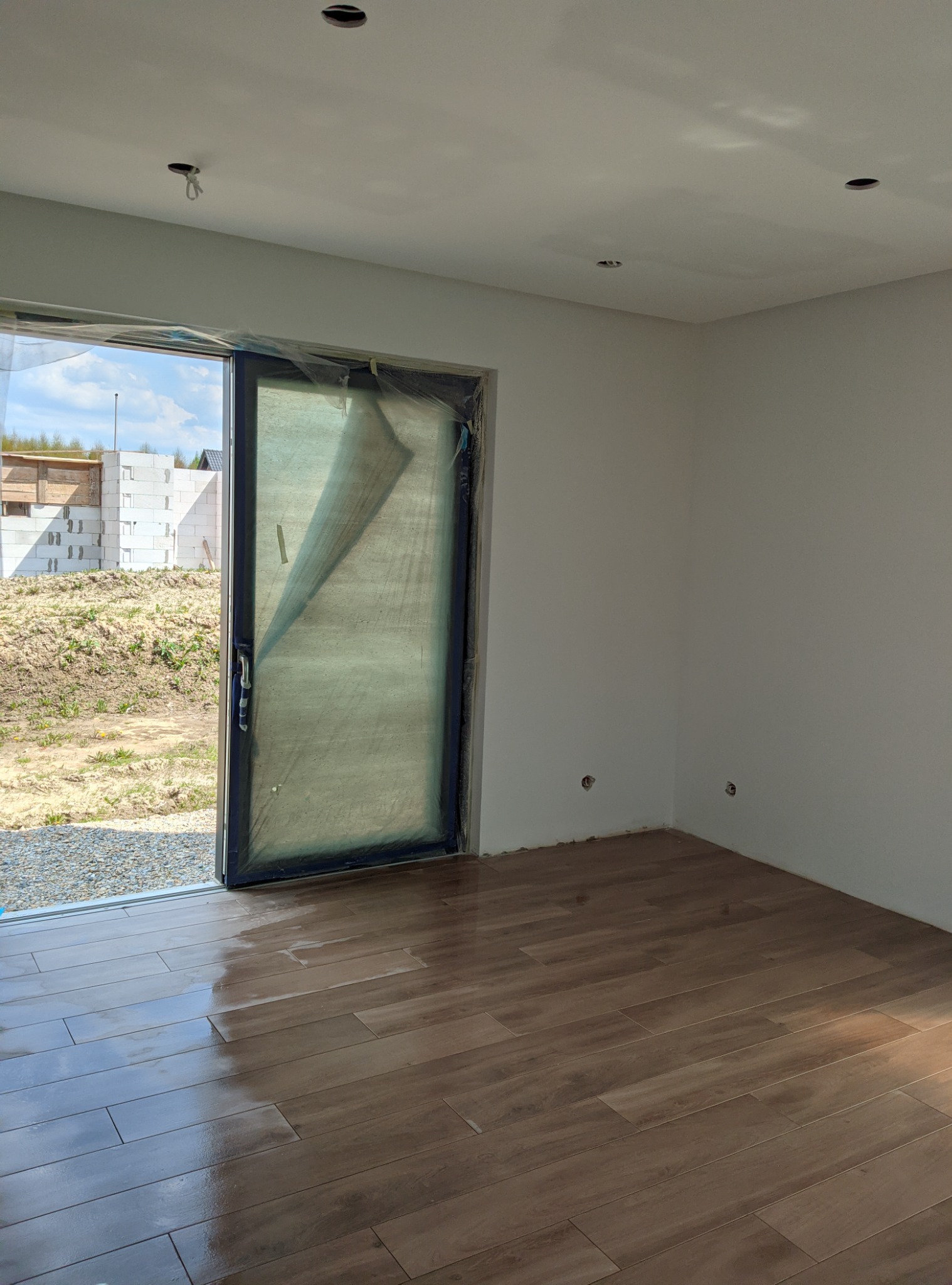 Wnętrze w trakcie prac wykończeniowych: świeżo położona podłoga z płytek imitujących drewno, ściany przygotowane pod malowanie, zabezpieczone folią drzwi balkonowe, widoczne otwory w suficie...