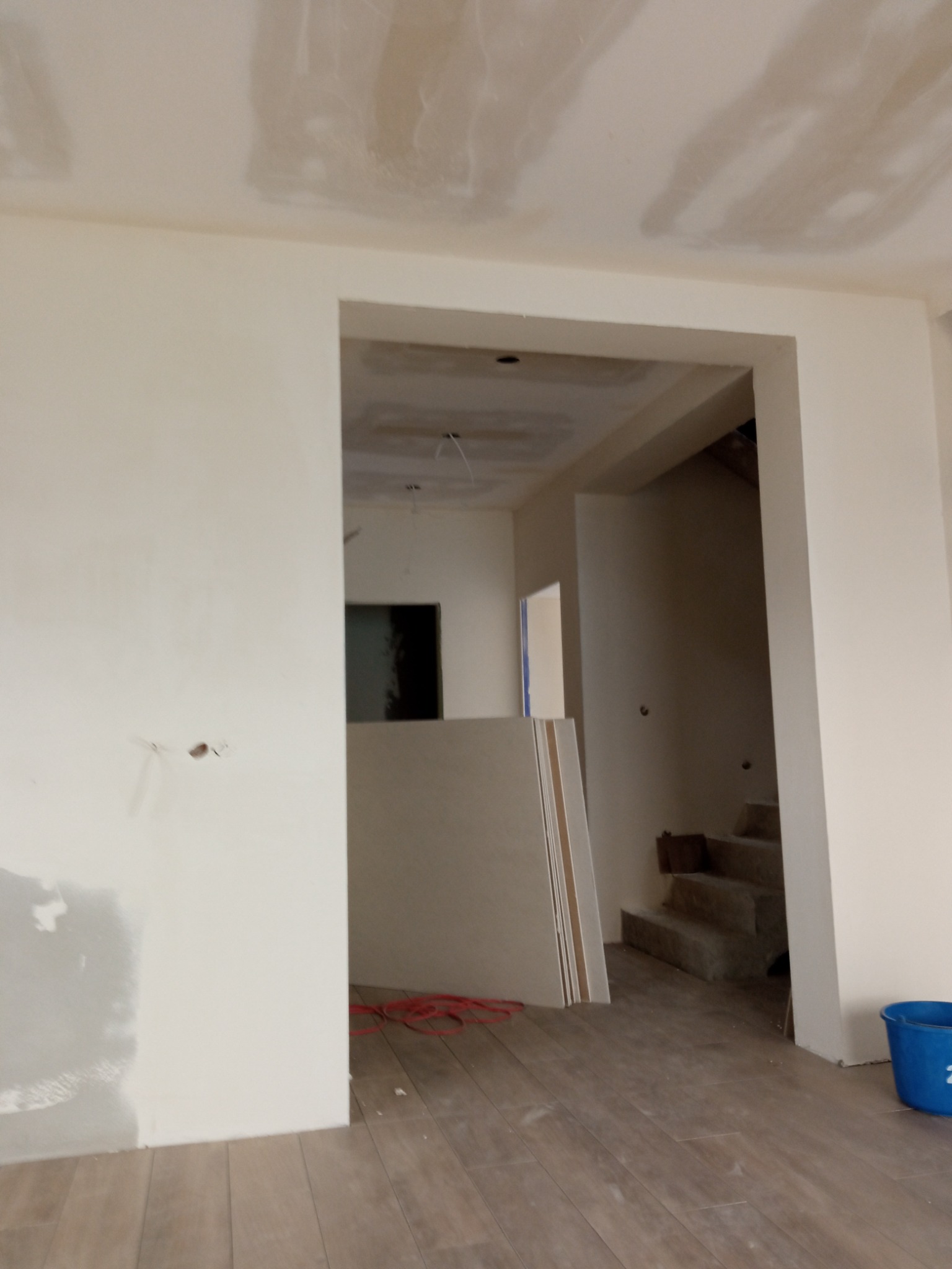 Wnętrze w trakcie remontu: ściany częściowo pokryte gładzią, widoczne płyty kartonowo-gipsowe oparte o ścianę, drewnopodobna podłoga i niebieskie wiadro.