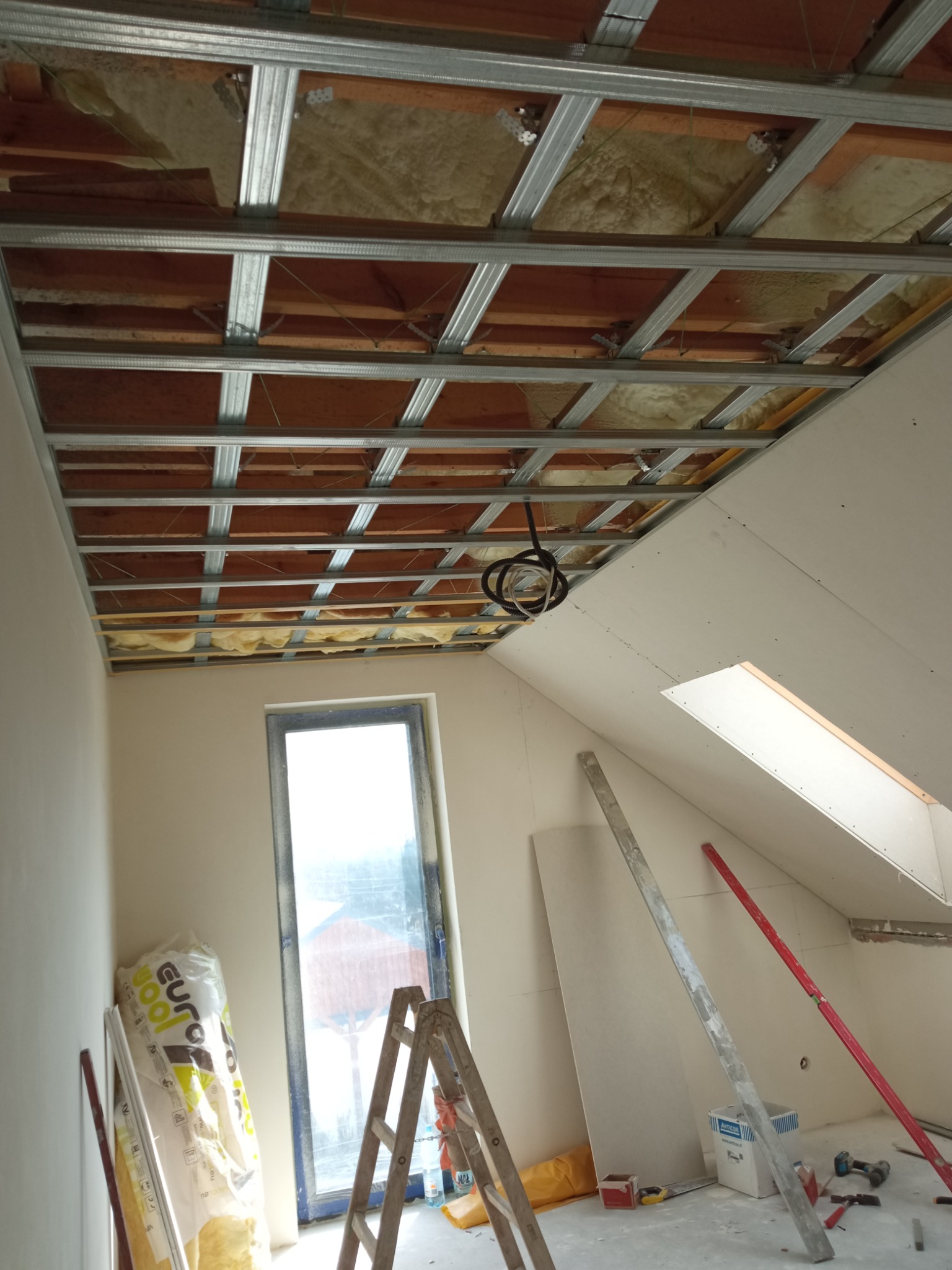 Montaż sufitu podwieszanego z płyt karton gips w poddaszu, widoczna metalowa konstrukcja, ocieplenie wełną mineralną, okno dachowe, drabina i materiały budowlane