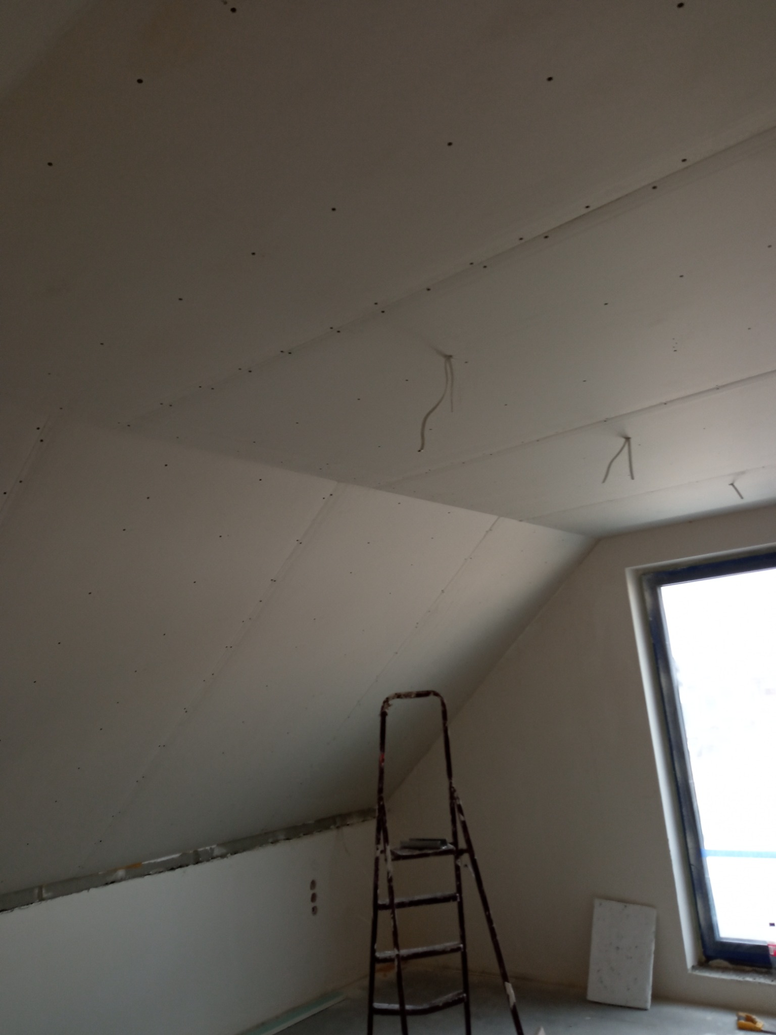 Montaż płyt karton gips na skośnym poddaszu, widoczne wkręty, wystające kable, drabina i okno dachowe.