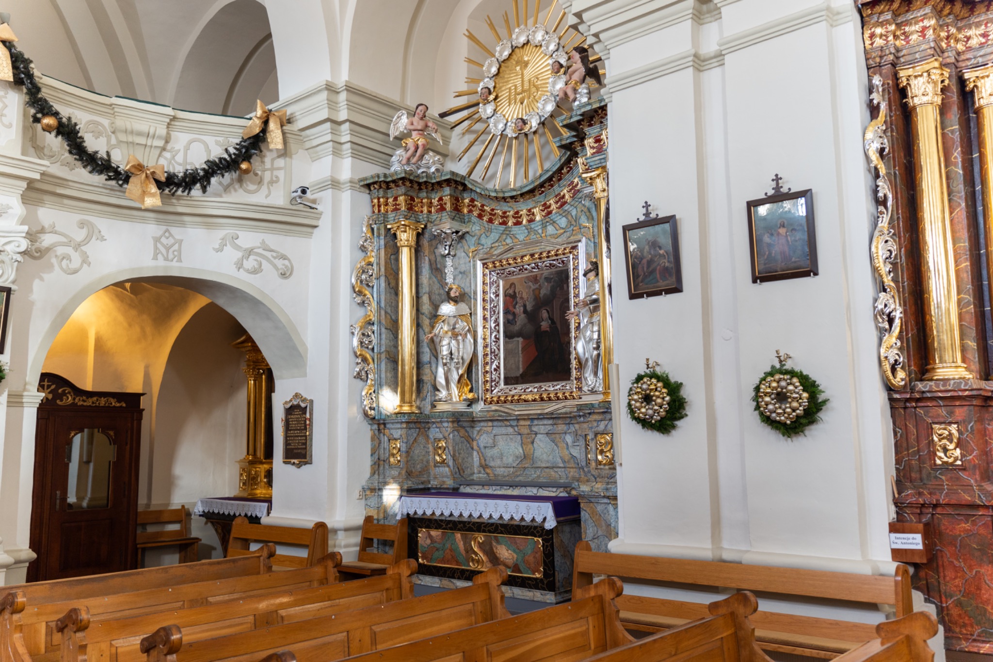 Wnętrze kościoła z barokowym ołtarzem głównym zdobionym figurami aniołów i złoceniami, drewniane ławki w pierwszym planie oraz dekoracje świąteczne.