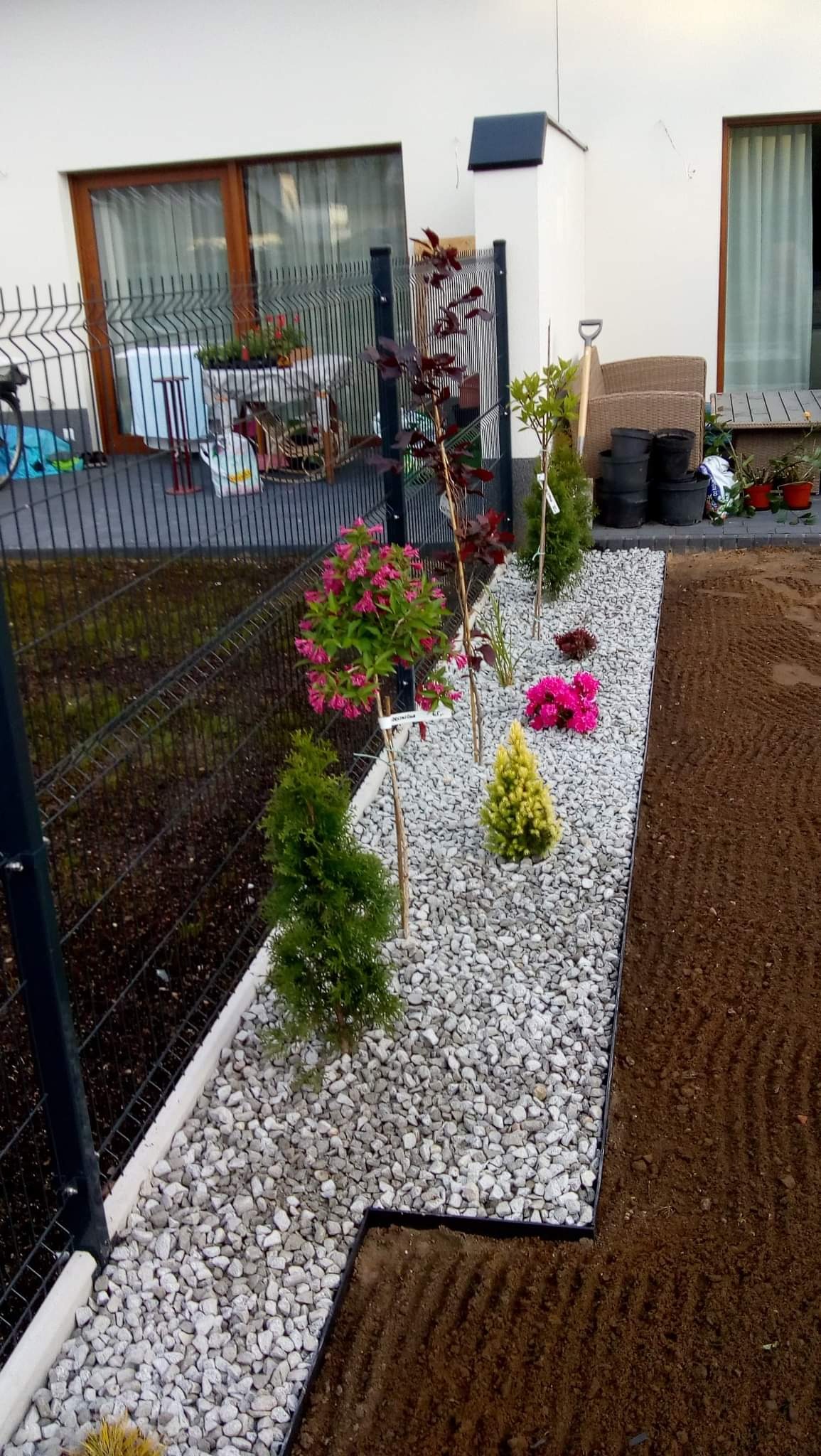 Świeżo posadzone drzewka i krzewy ozdobne, podsypane białym grysem, oddzielone obrzeżem od przygotowanej pod trawnik ziemi, widok z góry.